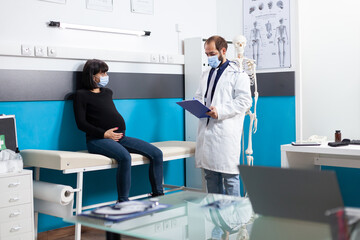 Specialist with face mask consulting pregnant woman in medical cabinet. Patient expecting child and doctor meeting at checkup visit during coronavirus pandemic. Pregnancy examination