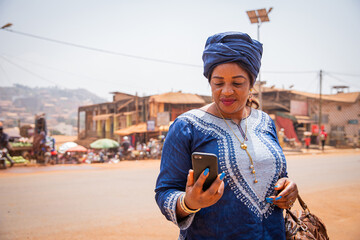 An African woman dressed in a traditional dress looks at the screen of her smartphone. 50-year-old...