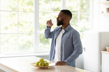 Calm millennial african american bearded man with closed eyes drinks water from glass in kitchen...