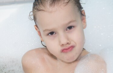 Children's hygiene. A cheerful little girl with foam in the bathroom looks out of the bathtub with an important facial expression and pouting lips.