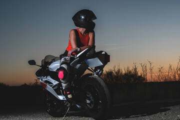 young biker woman with safety helmet sitting on her motorcycle outdoors in the field during a sunset