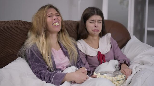 Two young absorbed women watching movie on TV start crying sympathizing actors. Portrait of Caucasian millennial ladies resting indoors on weekend enjoying romantic film