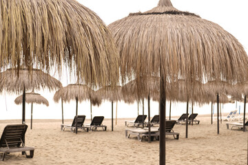Typical straw beach umbrella in province of Malaga, Andalusia, Spain. Straw beach umbrellas in a row, Mediterranean coast, sandy beach. Cloudy day, fog on the sea