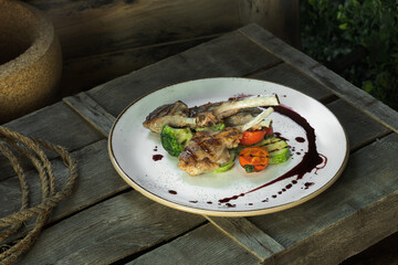 grilled rack of lamb and garnished with drops of sauce lies on a white dish along with tomatoes, zucchini and broccoli. The dish stands on a wooden box, rustic style
