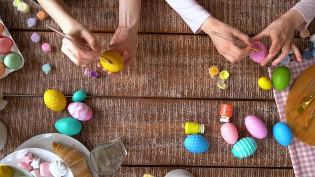 Happy Easter atmosphere! Close up funny, pretty, creative mom teacher, explain, to her cute, small, joyful daughter child how to draw, paint, decorate Easter eggs, together, sitting at desk