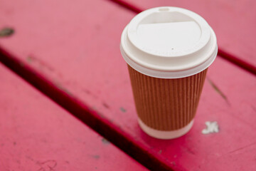 Cardboard cup with plastic lid for coffee or tee on a wooden red bench in a park. Outdoor refreshment concept. Selective focus.