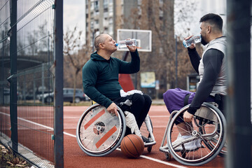 Wheelchair-bound basketball players drink water after practicing on outdoor court.