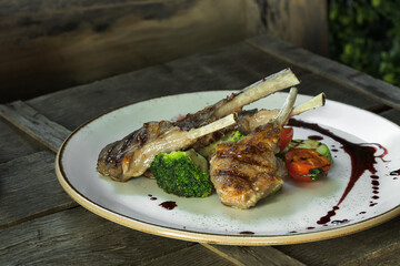 grilled rack of lamb and garnished with drops of sauce lies on a white dish along with tomatoes, zucchini and broccoli. The dish stands on a wooden box, rustic style, close-up