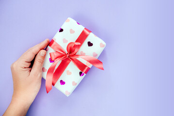A hand holding gift box wrapped in heart pattern paper with red ribbon bow on purple background