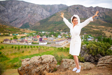 A girl in a terry, bathrobe with a towel rejoices and relaxes against the background of nature. Spa in a mountain resort. Rest in a sanatorium in the mountains. Purity and freshness of nature.