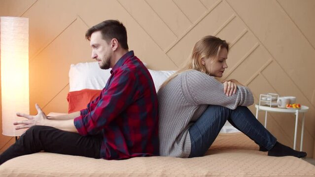 Husband and wife are sitting with their backs to each other on bed offended after quarrel, side view. Married couple is sitting with their backs to each other after squabble. Concept of resentment