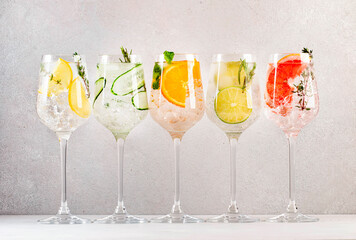 Gin and tonic cocktails set. Trendy Alcoholic drinks with lime, lemon, grapefruit, orange, cucumber, soda and spicy herbs in wine glasses, gray background. Summer cocktail party - obrazy, fototapety, plakaty