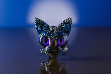 handmade craft ceramic brooch in the form of a muzzle of a cat with large blue glass eyes stands on a mirror surface, blue background