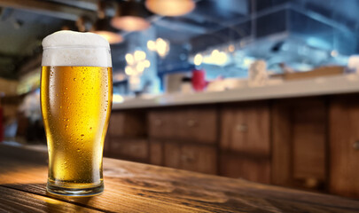 Cooled glass of beer on the wooden table. Blurred bar interior at the background.