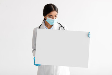 Young indian woman doctor in uniform, gloves and protective mask shows banner with blank space