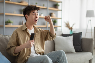 Excited asian guy spending weekend watching tv shaking clenched fist