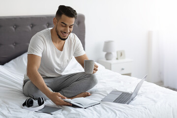 Remote Work. Arab Freelancer Man Working With Laptop And Notepad In Bed