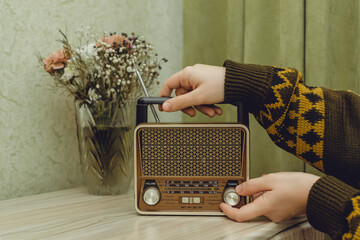 In a light green interior, the hands of a man in a knitted sweater with a pattern put a radio receiver on a chest of drawers and switches a musical composition, a bouquet of flowers stands behind. - obrazy, fototapety, plakaty
