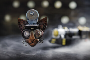 handmade craft ceramic brooch in the form of a muzzle of a steampunk driver's cat with a large glass eye levitates over a stone surface, against the background of a train in smoke, close-up - obrazy, fototapety, plakaty