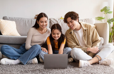 Smiling young asian female, male and teen girl sitting on floor and watching video, searching, browsing on laptop