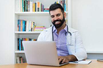 Laughing latin american mature doctor with hipster beard working at computer