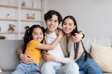 Cheerful asian couple and daughter hugs sits on sofa in living room interior