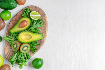 Top view of different green ripe vegetables and fruits sliced and whole such as avocado, apple, lime, lettuce and kiwi laying on brown cutting board on white wooden background with copy space