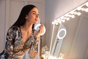 Young woman applying make up near illuminated mirror indoors