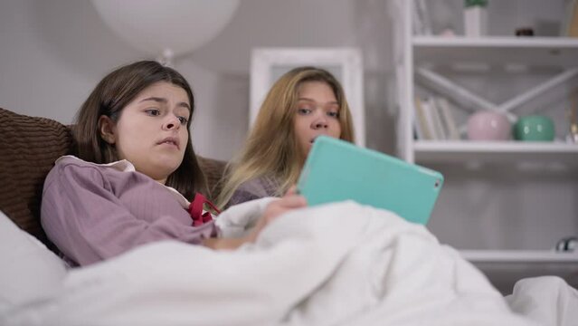 Scared young women with frightened facial expression watching horror movie online on tablet at pajama party. Portrait of Caucasian friends leaning back sitting on couch shocked with film