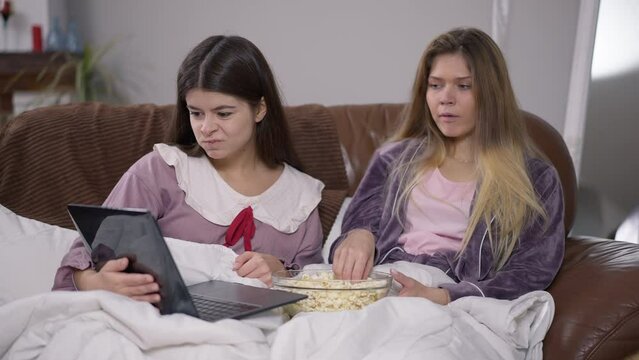 Two young absorbed women watching film online on laptop eating popcorn talking. Relaxed carefree Caucasian friends in pajamas enjoying movie on weekend indoors