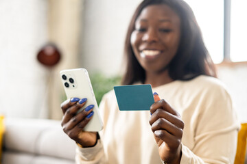 Selective focus at banking card and smartphone in hands of smiling african-american woman, happy lady using mobile app for ordering food delivery, shopping online, making money transaction. E-banking