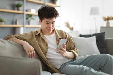 Smiling asian man using smartphone at home sitting on couch