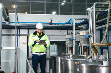 Caucasian technician engineer man in uniform with tablet checking and control boiler tanks and liquid pipeline in production line at factory