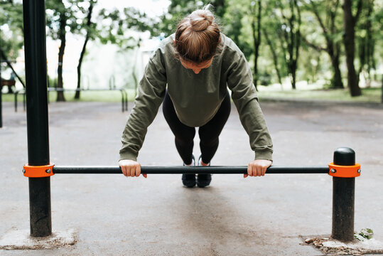 Athletic young woman doing push-ups on horizontal bar, physical exercises, strength training, fitness in sports ground. Front view of slim sportswoman exercising body in park outdoors. Selective focus