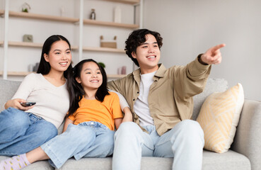 Smiling japanese young man shows on empty space with hand to wife and teen girl, sit on sofa in living room