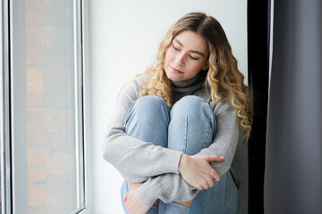 Woman in grey sweater and blue jeans sits on windowsill, watching in the window. Depressed girl thinking at the window