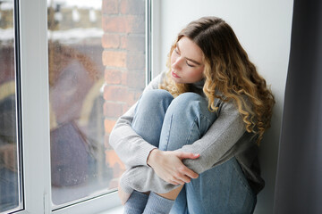 Woman in grey sweater and blue jeans sits on windowsill, watching in the window. Depressed girl thinking at the window