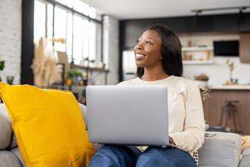 Inspired smiling african-american woman using laptop sitting down on comfortable couch, businesswoman answering emails. Ethnic female freelancer typing on the keyboard, studying or working on distance