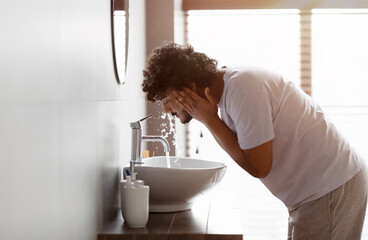 Young indian man washing face with clear water from tap bending over sink, doing skincare facial routine in bathroom - Powered by Adobe