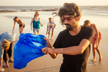 handsome indian man in the foreground collecting plastic from sand into the bag , his mixed race...