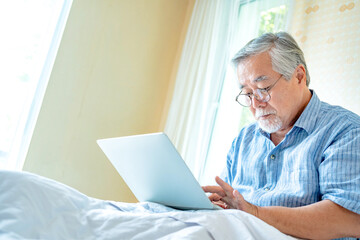 Happy old man positive, Portrait Asian Senior man and eyeglasses using computer laptop in bedroom at home, Old elderly male Technology communication retirement lifestyles concept.