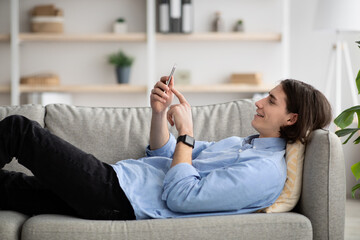 Man enjoying spare time alone lying on sofa and watching videos on cellphone, relaxing at home, using tech