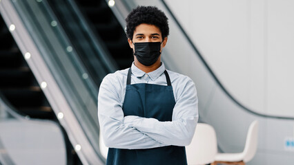 African american man guy restaurant cafe male worker in medical mask looking camera posing crossing arms waiter salesman wearing apron standing at work space covid pandemic quarantine. Small business
