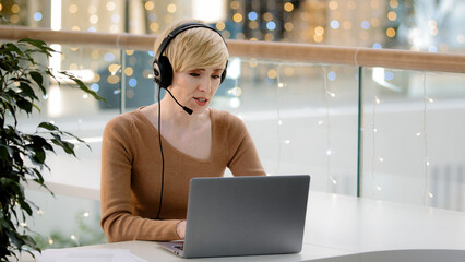 Middle aged adult 40s business woman in headset microphone talking at laptop web camera...