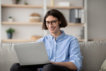 Fototapeta premium Remote work. Happy freelancer guy working with laptop at home, sitting on couch, enjoying distance job opportunities