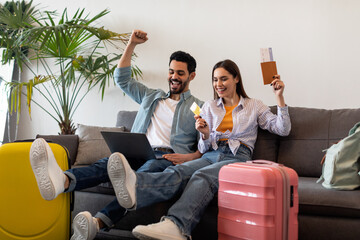 Happy couple booking hotel online using laptop and credit card, sitting with packed suitcases, passports and tickets