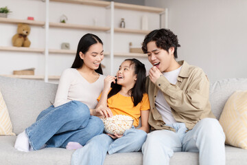 Satisfied young chinese woman, man and teenager girl eat popcorn, sit on sofa in living room interior and have fun