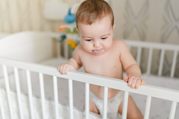 little european baby in a diaper stands in a crib at home, baby sticking out tongue