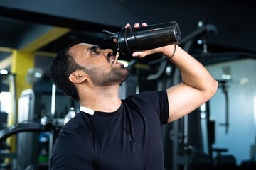 bodybuilder drinking or taking protein powder and mixing with water on bottle by shaking at gym -...