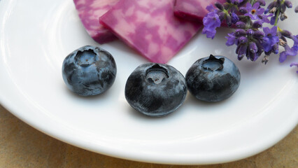 three blueberries on a white plate on a blurred background of pieces of lilac lavender cheese with lavender inflorescences. selective focus